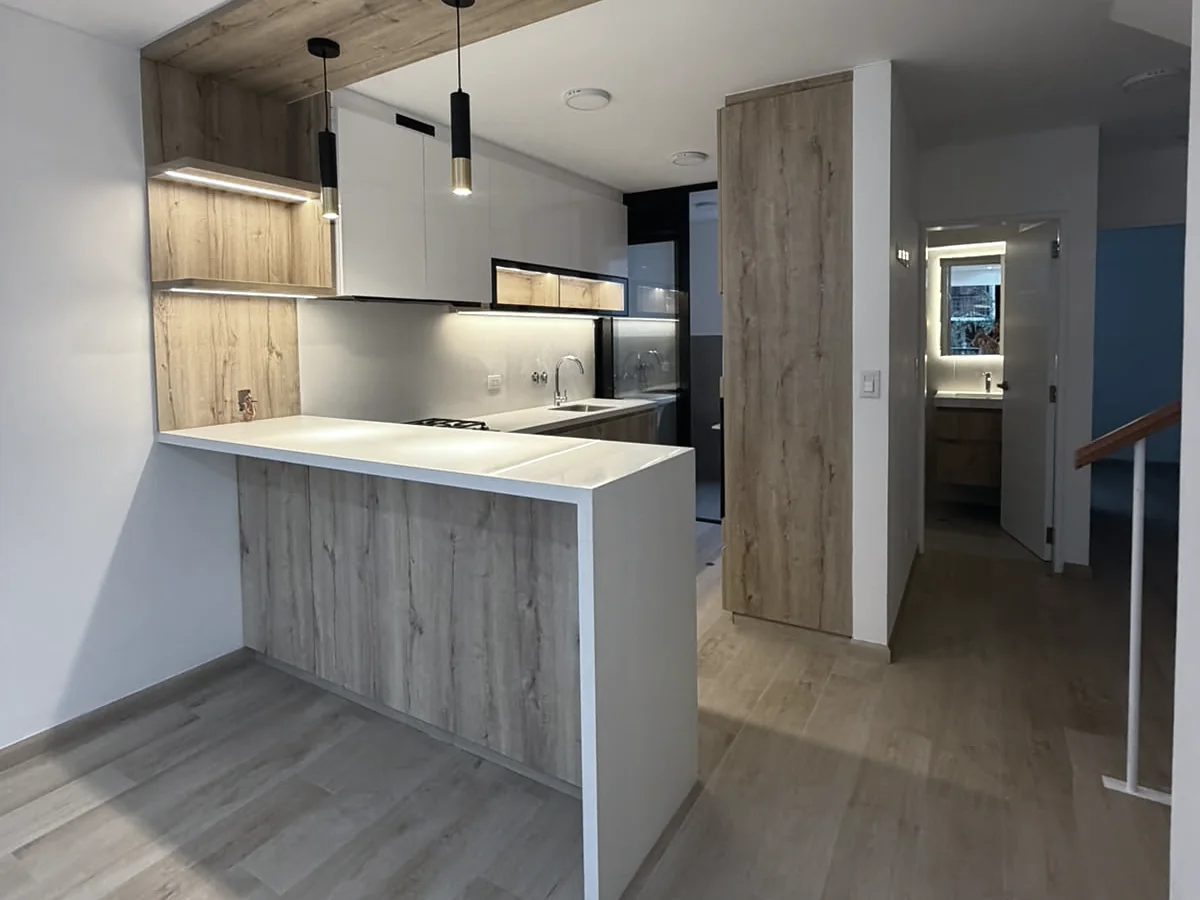 Cocina remodelada con barra blanca, muebles de madera y luces colgantes